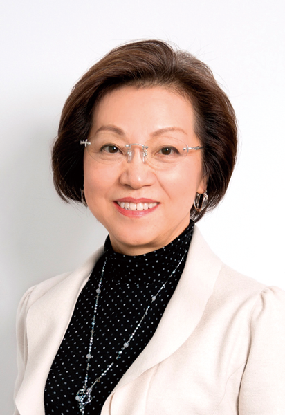 An older Japanese woman with a short bob wears glasses, earrings, and a necklace. She wears a black turtleneck with white polka dots and a white blazer.